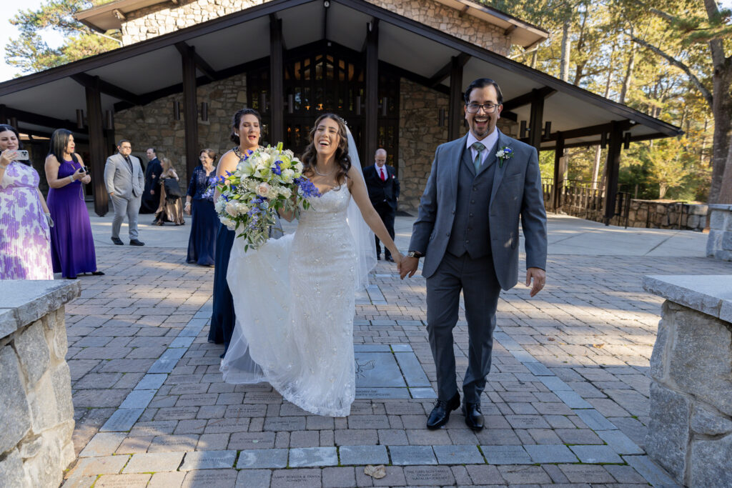 The bride and groom exit the wedding ceremony and smiling at the camera. Bride is holding her flower boutique.