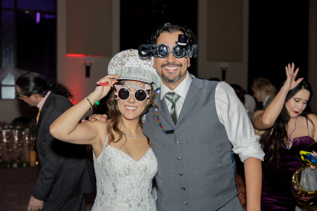 The bride and groom smiling at the camera during their wedding reception while they wear their custom and fun glasses and hats.