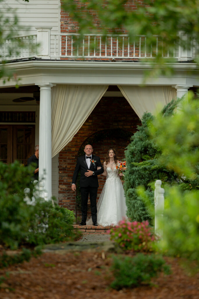 A photograph of the bride and her father, during the ceremony, standing at the edge of The Carl House, waiting to walk forward to the altar.