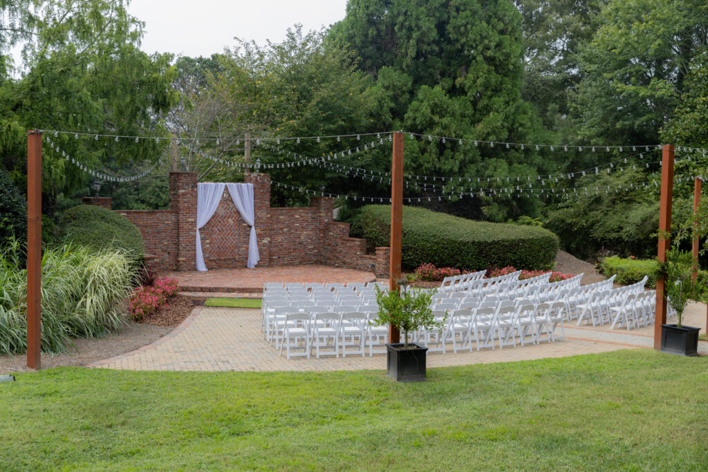 The wedding ceremony from afar, before anyone enters the frame. Full of luscious greenery, red brick presentation, and cute string lights.