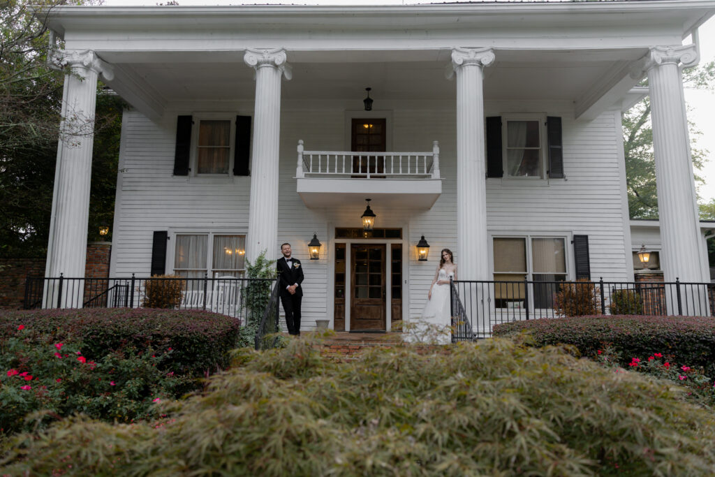 The bride and groom posing in front of the Carl House, one on each column pillar looking at the camera smiling.